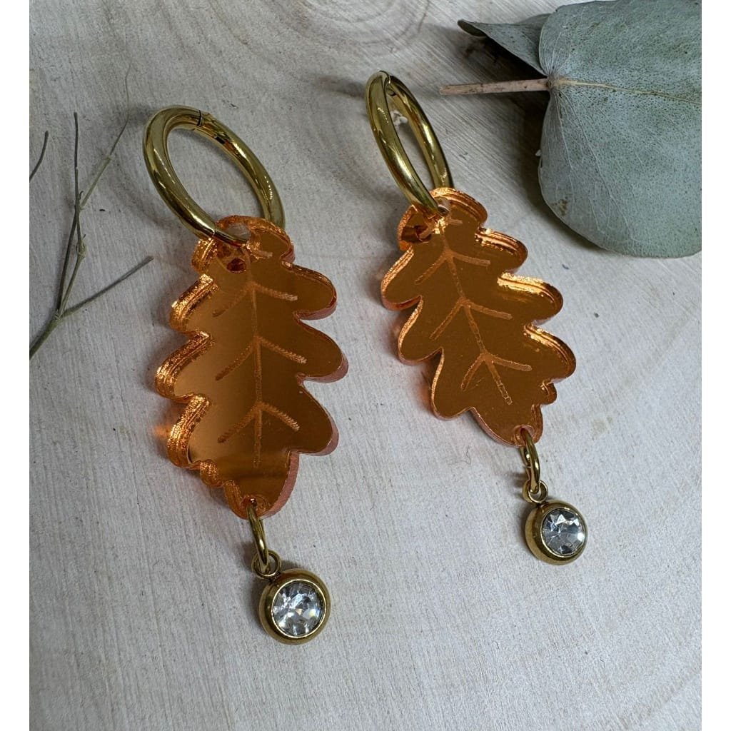 Los pendientes Hoja de Roble con piedra de cristal son una auténtica joya artesanal pensada para quienes buscan elegancia natural y un toque de brillo único. Fabricados en metacrilato efecto espejo, estos pendientes capturan la luz desde todos los ángulos, creando un reflejo que resalta la forma delicada de la hoja y el brillo sutil de la piedra central.