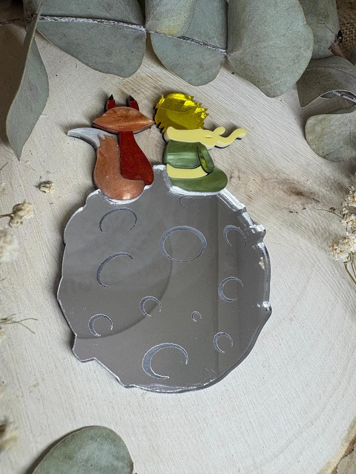 El Broche El Principito es una pieza única que combina artesanía, emoción y diseño. Inspirado en uno de los personajes más entrañables de la literatura universal, este broche está realizado a mano minuciosamente, cuidando cada detalle para crear una joya especial, delicada y llena de significado. Fabricado con metacrilatos de importación de alta calidad, incorpora acabados efecto espejo y marmoleados, que aportan profundidad, brillo y un juego de luces espectacular. Cada capa ha sido cortada con máquina láser de alta precisión, lo que permite un encaje perfecto y una definición impecable en cada forma. Su diseño representa al Principito junto a su inseparable zorro sobre su pequeño planeta, una escena que transmite ternura, amistad y magia. Gracias a la combinación de colores y materiales, el broche resulta llamativo sin perder elegancia, convirtiéndose en un complemento ideal tanto para el día a día como para ocasiones especiales.