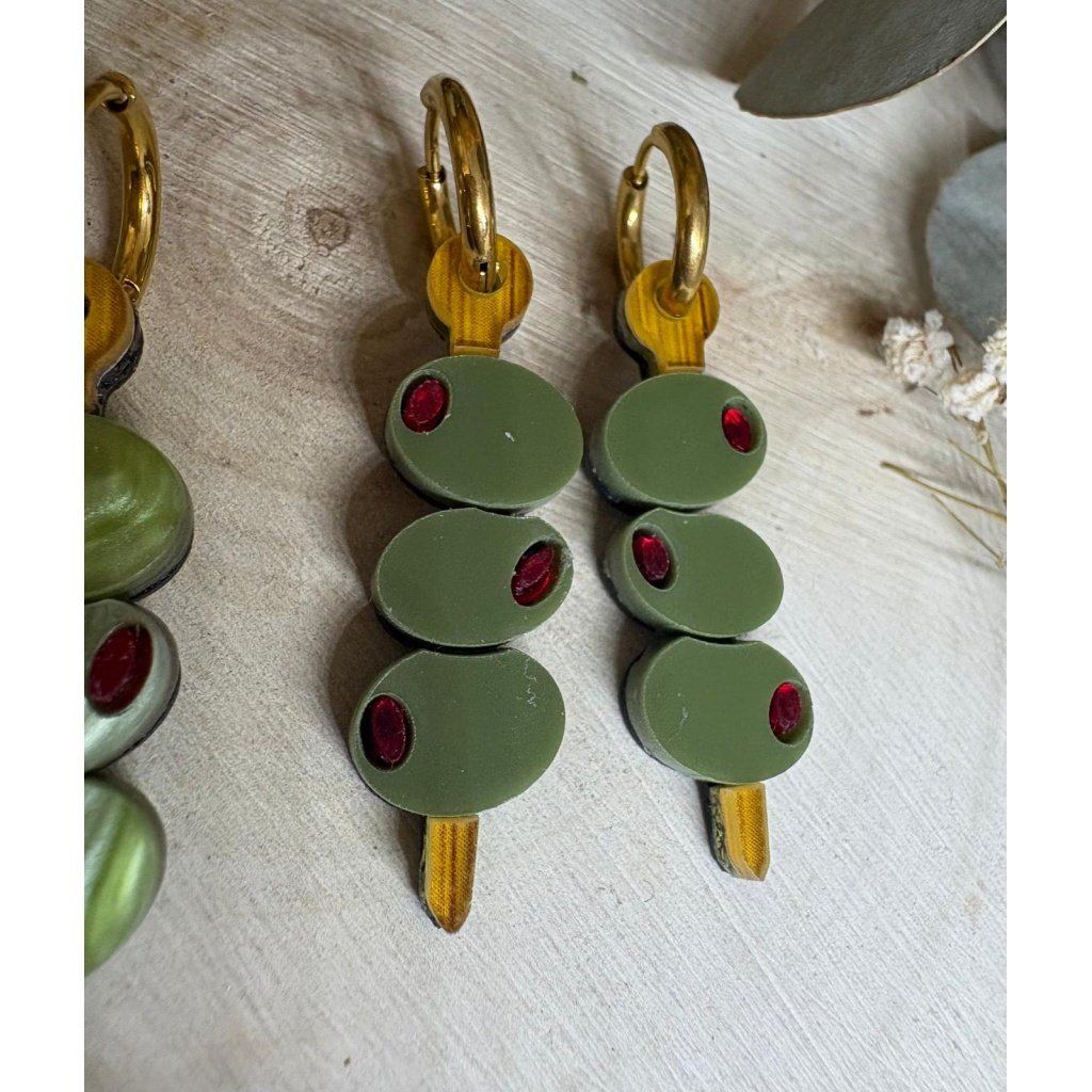 Pendientes Banderilla de Aceitunas  Los Pendientes Banderilla de Aceitunas son el accesorio perfecto para quienes buscan un toque divertido, original y con mucha personalidad en sus looks. Inspirados en uno de los aperitivos más icónicos, estos pendientes reinterpretan la clásica banderilla en una pieza única de joyería que no pasa desapercibida. Este modelo exclusivo de JudithconAlma está diseñado para aportar frescura y estilo, evocando ese ambiente veraniego de terrazas, tapeo y momentos especiales. Son ideales para completar looks desenfadados, dar un toque diferente a conjuntos básicos o incluso como pieza protagonista en outfits más atrevidos. Pendientes Banderilla de Aceitunas – Diseño exclusivo JudithconAlma Están elaborados con metacrilato de alta calidad, combinando diferentes acabados que aportan riqueza visual y un acabado muy cuidado. Puedes encontrarlos en dos versiones: verde marmoleado, con un efecto más dinámico y lleno de matices, y verde liso, más uniforme y elegante. Ambos diseños incorporan detalles en metacrilato efecto madera, que recrean el palillo de la banderilla y aportan un contraste cálido y natural. Cada pendiente representa una pequeña brocheta de aceitunas, con detalles que imitan su forma y color, logrando un resultado creativo, divertido y muy reconocible. A pesar de su diseño llamativo, son ligeros y muy cómodos, perfectos para llevar durante todo el día sin molestias. Incorporan un aro de acero inoxidable, un material resistente, duradero y apto para pieles sensibles y personas con alergias, lo que garantiza una experiencia cómoda y segura. Su tamaño de 3,5 x 1 cm (sin aro) los hace ideales para destacar sin resultar excesivos. Los Pendientes Banderilla de Aceitunas son una apuesta segura si buscas pendientes originales, diferentes y con aire veraniego, perfectos para eventos, vacaciones o para darle un toque divertido a tu día a día. También son una opción ideal para regalar algo único y lleno de creatividad. Un diseño que refleja el espíritu de JudithconAlma: piezas exclusivas, con alma, que combinan originalidad, calidad y estilo propio.