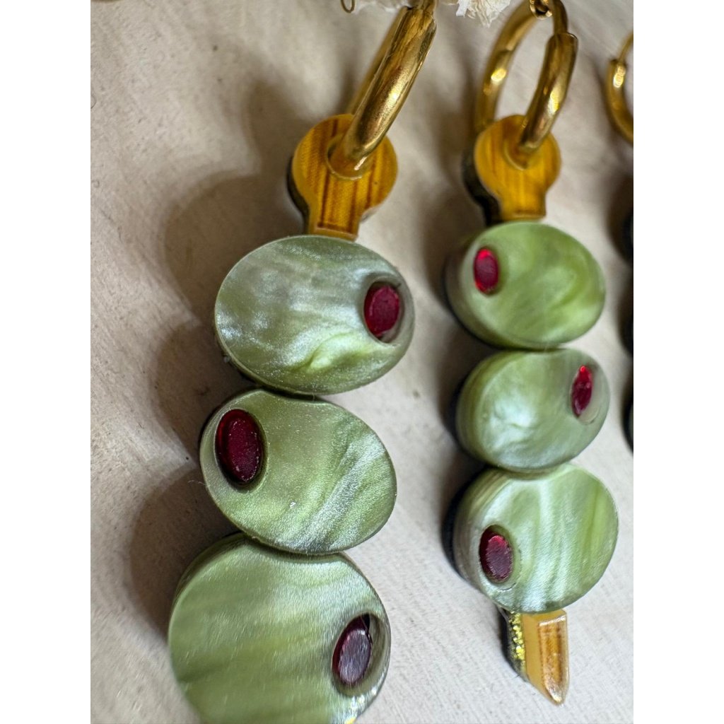 Pendientes Banderilla de Aceitunas  Los Pendientes Banderilla de Aceitunas son el accesorio perfecto para quienes buscan un toque divertido, original y con mucha personalidad en sus looks. Inspirados en uno de los aperitivos más icónicos, estos pendientes reinterpretan la clásica banderilla en una pieza única de joyería que no pasa desapercibida. Este modelo exclusivo de JudithconAlma está diseñado para aportar frescura y estilo, evocando ese ambiente veraniego de terrazas, tapeo y momentos especiales. Son ideales para completar looks desenfadados, dar un toque diferente a conjuntos básicos o incluso como pieza protagonista en outfits más atrevidos. Pendientes Banderilla de Aceitunas – Diseño exclusivo JudithconAlma Están elaborados con metacrilato de alta calidad, combinando diferentes acabados que aportan riqueza visual y un acabado muy cuidado. Puedes encontrarlos en dos versiones: verde marmoleado, con un efecto más dinámico y lleno de matices, y verde liso, más uniforme y elegante. Ambos diseños incorporan detalles en metacrilato efecto madera, que recrean el palillo de la banderilla y aportan un contraste cálido y natural. Cada pendiente representa una pequeña brocheta de aceitunas, con detalles que imitan su forma y color, logrando un resultado creativo, divertido y muy reconocible. A pesar de su diseño llamativo, son ligeros y muy cómodos, perfectos para llevar durante todo el día sin molestias. Incorporan un aro de acero inoxidable, un material resistente, duradero y apto para pieles sensibles y personas con alergias, lo que garantiza una experiencia cómoda y segura. Su tamaño de 3,5 x 1 cm (sin aro) los hace ideales para destacar sin resultar excesivos. Los Pendientes Banderilla de Aceitunas son una apuesta segura si buscas pendientes originales, diferentes y con aire veraniego, perfectos para eventos, vacaciones o para darle un toque divertido a tu día a día. También son una opción ideal para regalar algo único y lleno de creatividad. Un diseño que refleja el espíritu de JudithconAlma: piezas exclusivas, con alma, que combinan originalidad, calidad y estilo propio.