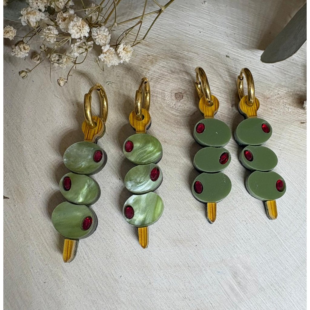 Pendientes Banderilla de Aceitunas  Los Pendientes Banderilla de Aceitunas son el accesorio perfecto para quienes buscan un toque divertido, original y con mucha personalidad en sus looks. Inspirados en uno de los aperitivos más icónicos, estos pendientes reinterpretan la clásica banderilla en una pieza única de joyería que no pasa desapercibida. Este modelo exclusivo de JudithconAlma está diseñado para aportar frescura y estilo, evocando ese ambiente veraniego de terrazas, tapeo y momentos especiales. Son ideales para completar looks desenfadados, dar un toque diferente a conjuntos básicos o incluso como pieza protagonista en outfits más atrevidos. Pendientes Banderilla de Aceitunas – Diseño exclusivo JudithconAlma Están elaborados con metacrilato de alta calidad, combinando diferentes acabados que aportan riqueza visual y un acabado muy cuidado. Puedes encontrarlos en dos versiones: verde marmoleado, con un efecto más dinámico y lleno de matices, y verde liso, más uniforme y elegante. Ambos diseños incorporan detalles en metacrilato efecto madera, que recrean el palillo de la banderilla y aportan un contraste cálido y natural. Cada pendiente representa una pequeña brocheta de aceitunas, con detalles que imitan su forma y color, logrando un resultado creativo, divertido y muy reconocible. A pesar de su diseño llamativo, son ligeros y muy cómodos, perfectos para llevar durante todo el día sin molestias. Incorporan un aro de acero inoxidable, un material resistente, duradero y apto para pieles sensibles y personas con alergias, lo que garantiza una experiencia cómoda y segura. Su tamaño de 3,5 x 1 cm (sin aro) los hace ideales para destacar sin resultar excesivos. Los Pendientes Banderilla de Aceitunas son una apuesta segura si buscas pendientes originales, diferentes y con aire veraniego, perfectos para eventos, vacaciones o para darle un toque divertido a tu día a día. También son una opción ideal para regalar algo único y lleno de creatividad. Un diseño que refleja el espíritu de JudithconAlma: piezas exclusivas, con alma, que combinan originalidad, calidad y estilo propio.