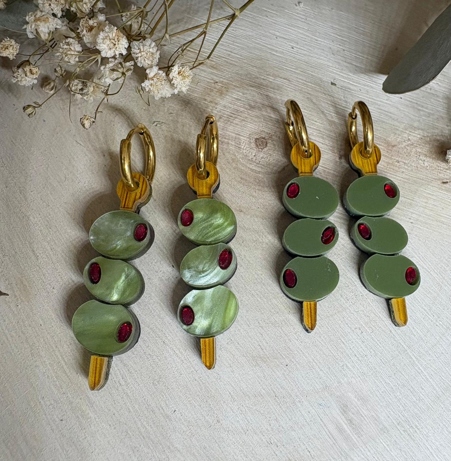 Pendientes Banderilla de Aceitunas  Los Pendientes Banderilla de Aceitunas son el accesorio perfecto para quienes buscan un toque divertido, original y con mucha personalidad en sus looks. Inspirados en uno de los aperitivos más icónicos, estos pendientes reinterpretan la clásica banderilla en una pieza única de joyería que no pasa desapercibida. Este modelo exclusivo de JudithconAlma está diseñado para aportar frescura y estilo, evocando ese ambiente veraniego de terrazas, tapeo y momentos especiales. Son ideales para completar looks desenfadados, dar un toque diferente a conjuntos básicos o incluso como pieza protagonista en outfits más atrevidos. Pendientes Banderilla de Aceitunas – Diseño exclusivo JudithconAlma Están elaborados con metacrilato de alta calidad, combinando diferentes acabados que aportan riqueza visual y un acabado muy cuidado. Puedes encontrarlos en dos versiones: verde marmoleado, con un efecto más dinámico y lleno de matices, y verde liso, más uniforme y elegante. Ambos diseños incorporan detalles en metacrilato efecto madera, que recrean el palillo de la banderilla y aportan un contraste cálido y natural. Cada pendiente representa una pequeña brocheta de aceitunas, con detalles que imitan su forma y color, logrando un resultado creativo, divertido y muy reconocible. A pesar de su diseño llamativo, son ligeros y muy cómodos, perfectos para llevar durante todo el día sin molestias. Incorporan un aro de acero inoxidable, un material resistente, duradero y apto para pieles sensibles y personas con alergias, lo que garantiza una experiencia cómoda y segura. Su tamaño de 3,5 x 1 cm (sin aro) los hace ideales para destacar sin resultar excesivos. Los Pendientes Banderilla de Aceitunas son una apuesta segura si buscas pendientes originales, diferentes y con aire veraniego, perfectos para eventos, vacaciones o para darle un toque divertido a tu día a día. También son una opción ideal para regalar algo único y lleno de creatividad. Un diseño que refleja el espíritu de JudithconAlma: piezas exclusivas, con alma, que combinan originalidad, calidad y estilo propio.